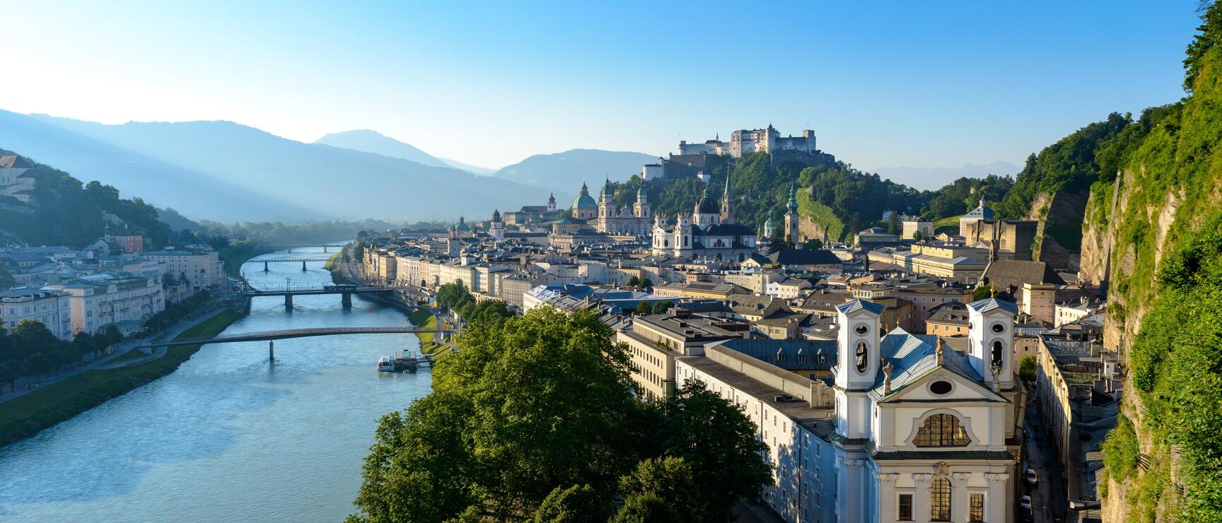 Panoramablick auf Salzburgs Altstadt mit Salzach, Festung Hohensalzburg auf dem Felsen und Alpen im Hintergrund.