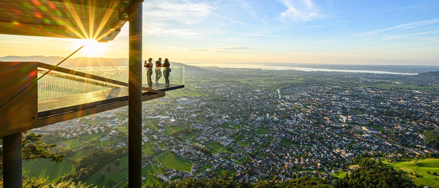 Drei Personen auf einer Aussichtsplattform über Dornbirn, Bodensee im Hintergrund, Sonnenuntergang.