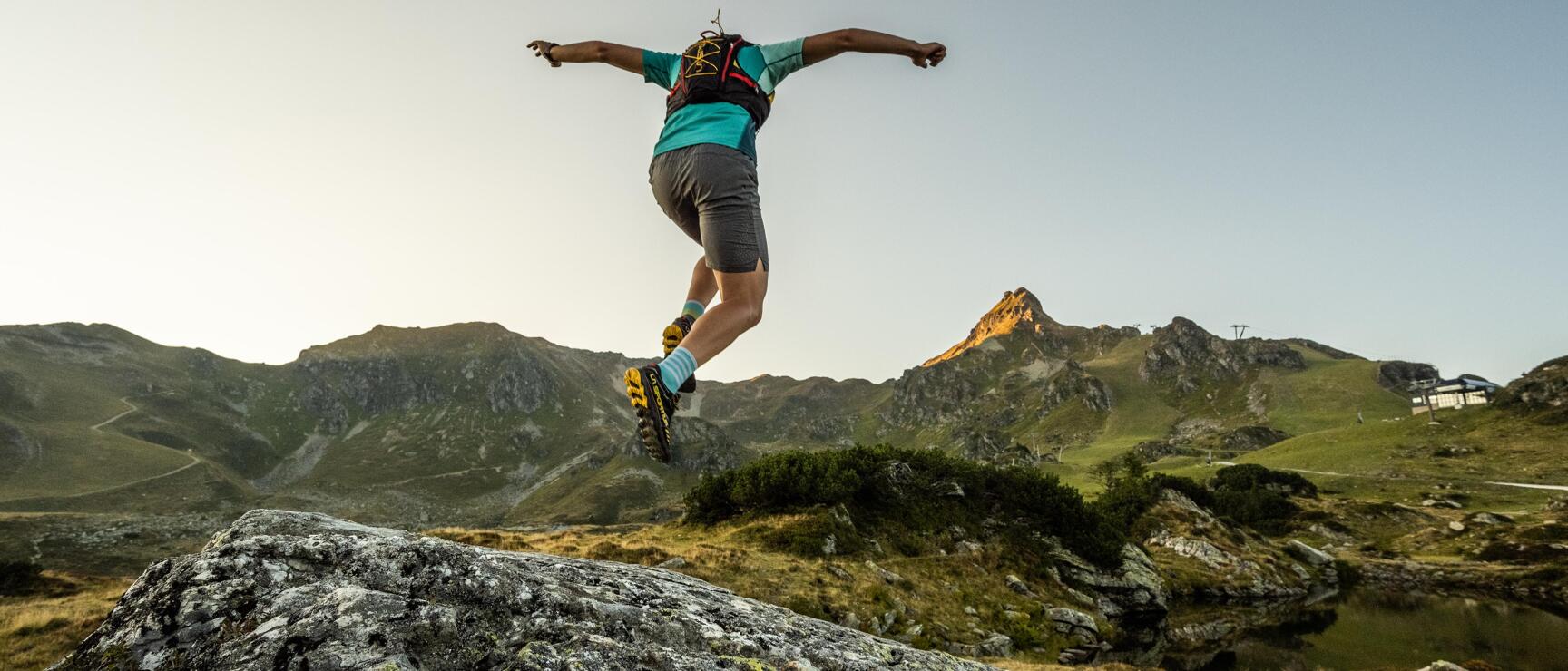 Person with backpack jumping off a rock, mountain panorama with alpine meadows in the background.