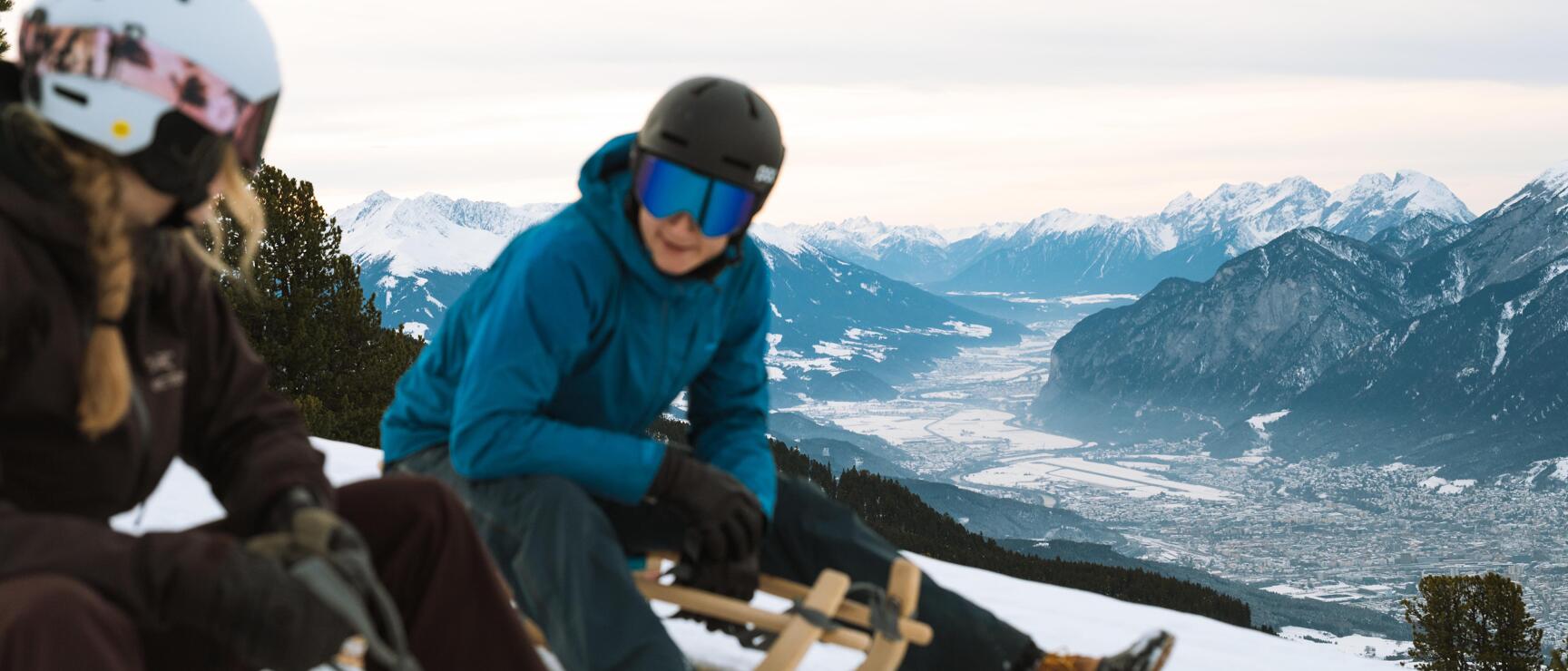 Two people with helmets on wooden sledge, snowy mountain landscape with Inn Valley in background.