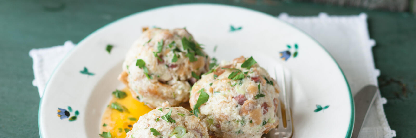 Four bread dumplings with parsley and butter sauce on white patterned plate with cutlery.