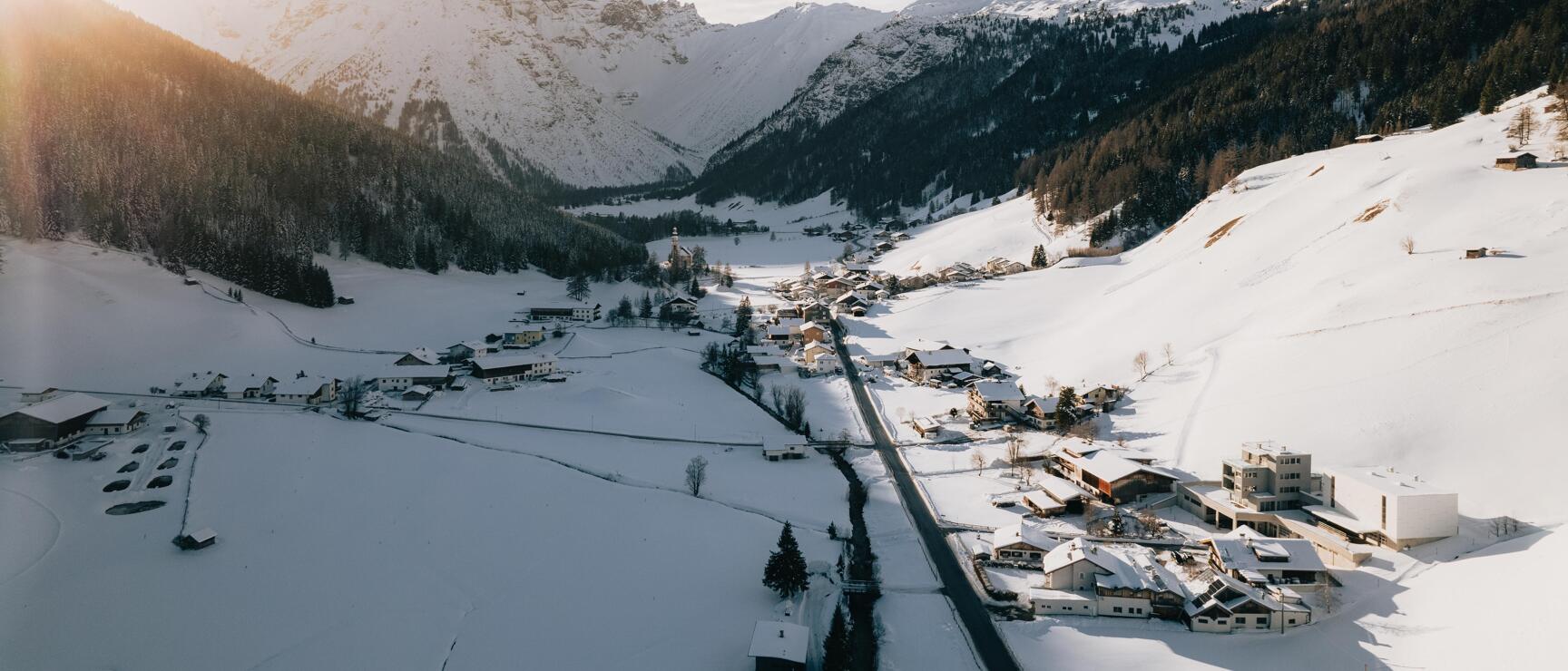 Aerial view of a snowy Alpine valley with village, road and snow-capped peaks in backlighting.