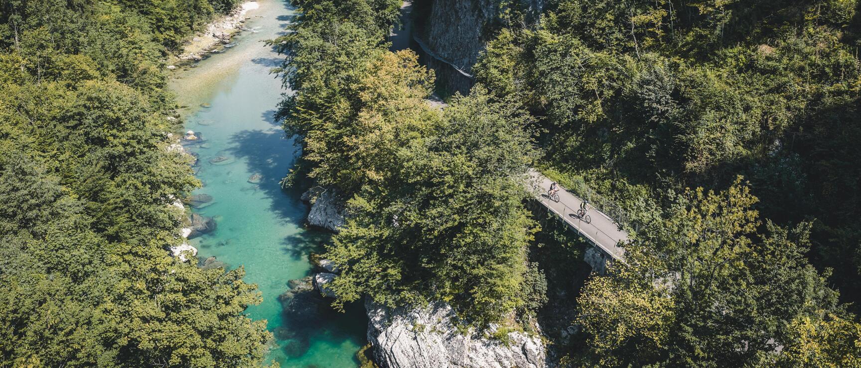 Luftaufnahme: Zwei Radfahrer auf Brücke über türkisfarbenem Fluss in bewaldeter Felsschlucht.