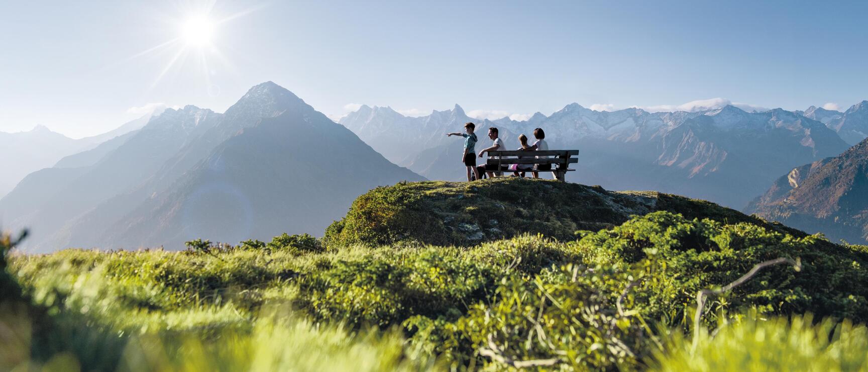 Vier Personen auf einer Bergbank, eine zeigt in die Ferne, Alpenpanorama mit Gipfeln im Hintergrund.