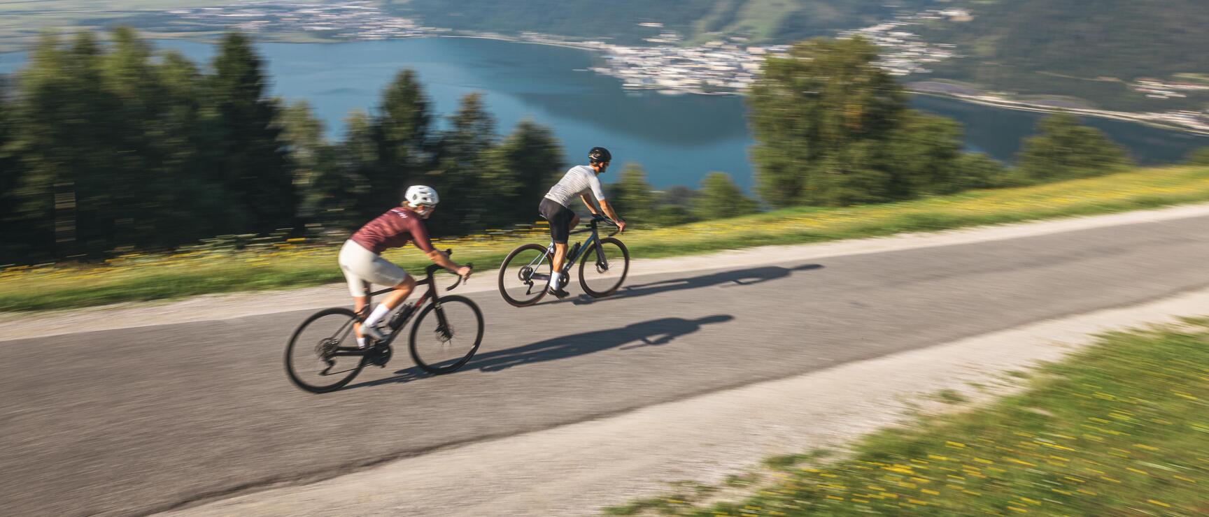 Two road cyclists on a mountain road, reservoir and Alpine panorama behind under blue sky.