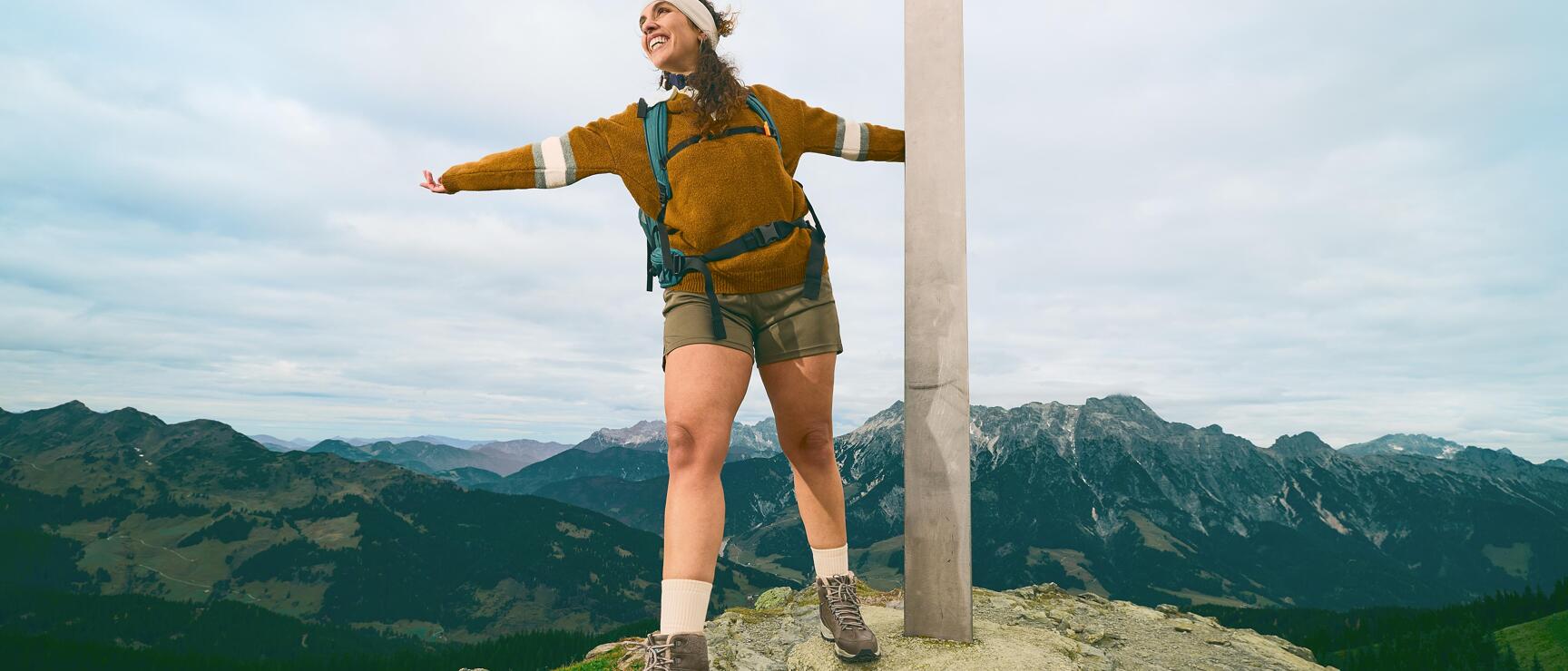 Woman with backpack standing at summit cross on rock, arms outstretched, mountain panorama in background