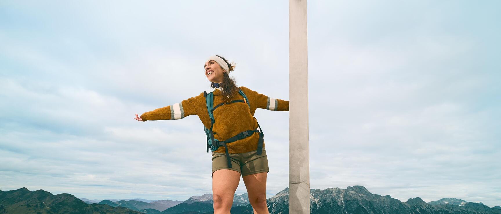 Woman with backpack standing at summit cross on rock, arms outstretched, mountain panorama in background