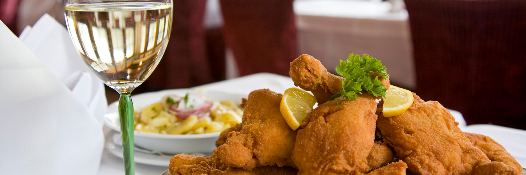 Fried chicken with lemon slices and parsley on plate, potato salad and white wine glass beside.