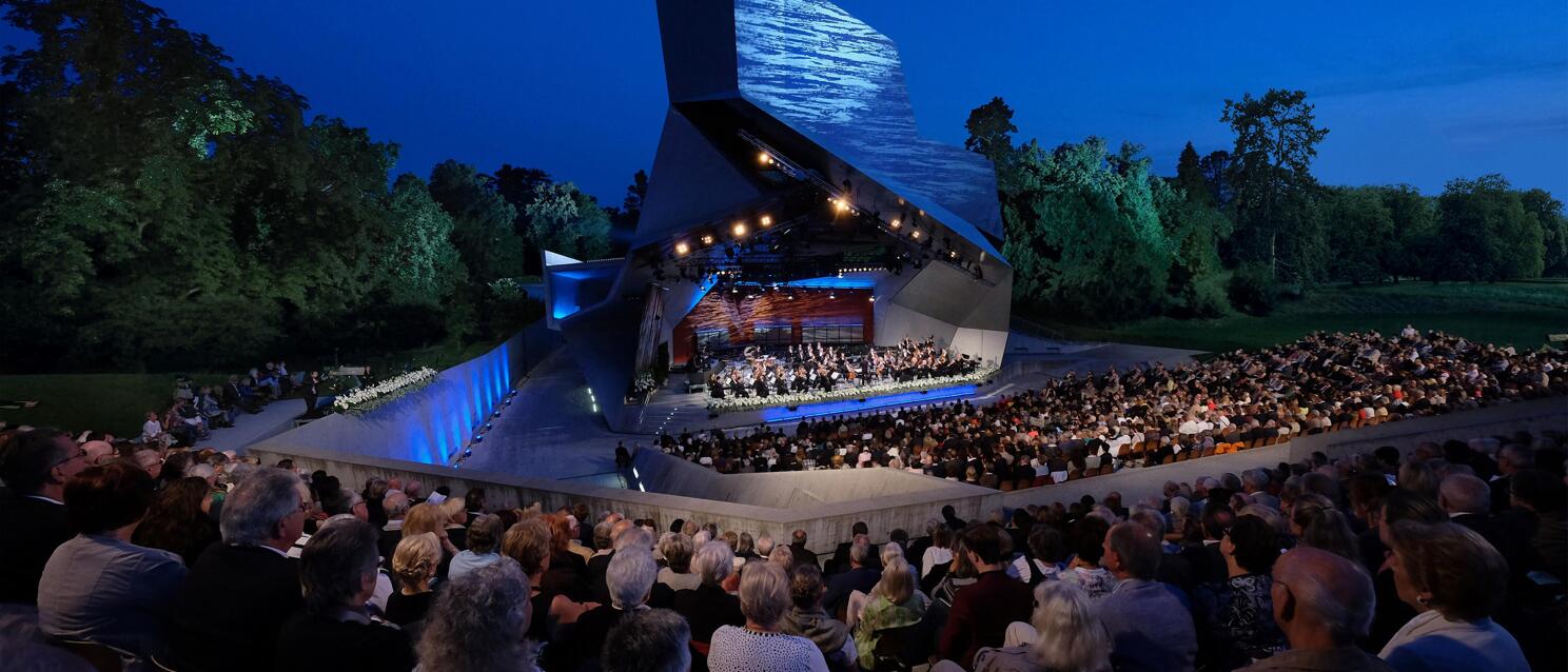 Fully booked open-air amphitheatre at night with orchestra on illuminated stage, modern stage roof, trees in background.