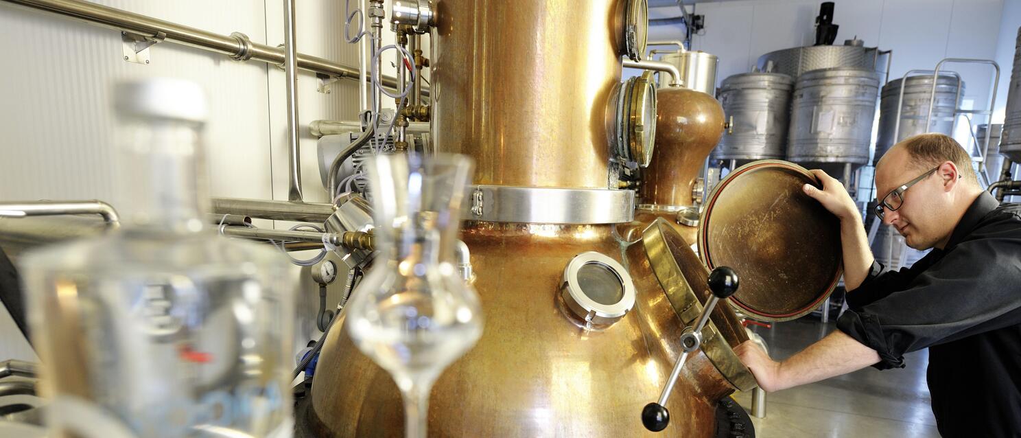 Man opens lid of copper distillery apparatus, schnapps glass and bottle in foreground.