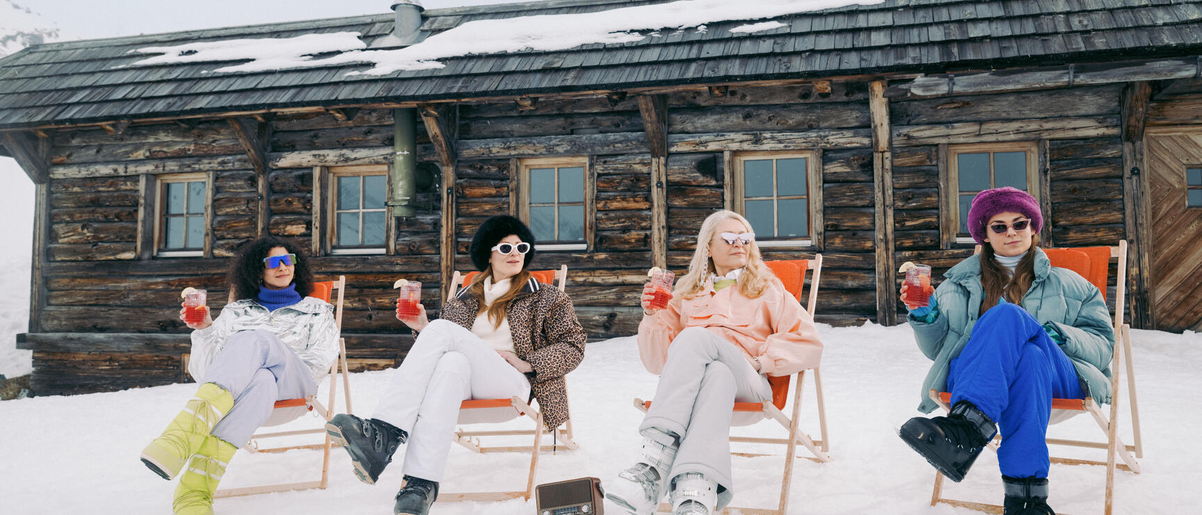Four women wearing sunglasses holding drinks in deckchairs outside a mountain hut in snow, mountains in background.