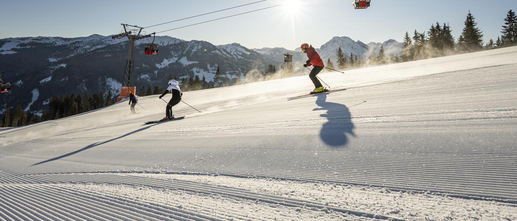 Two skiers on a groomed slope in sunshine, red gondola, snow-covered mountains in the background.