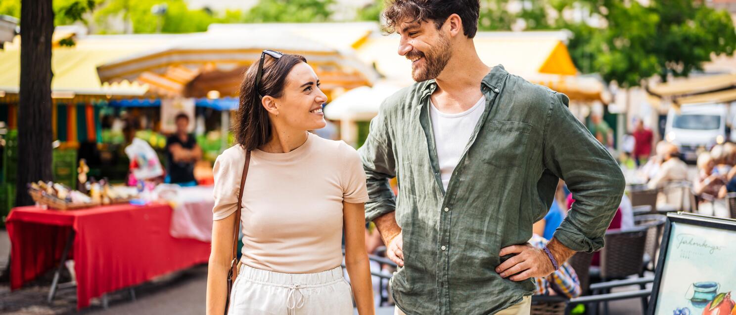 Smiling couple at weekly market with colourful market stalls and trees in background.