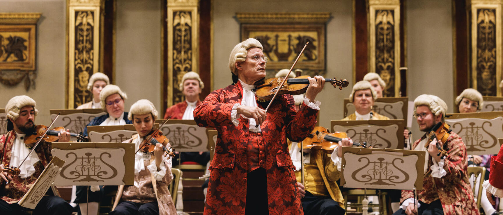 Violinist in red Baroque costume and wig playing violin with orchestra behind in Vienna Musikverein
