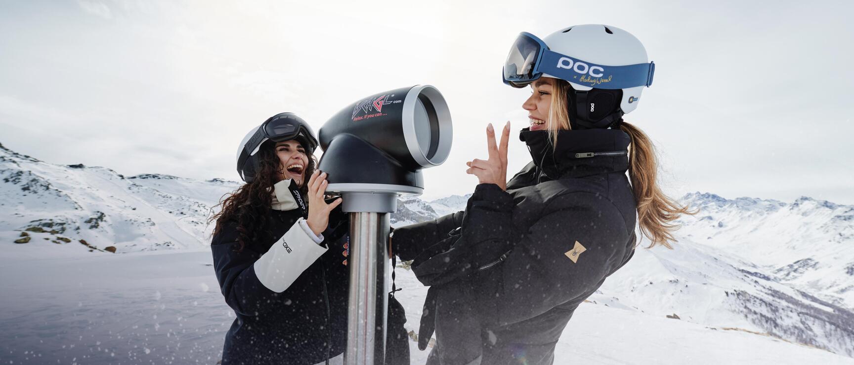 Two laughing women wearing ski helmets at a coin telescope on snowy mountain summit.