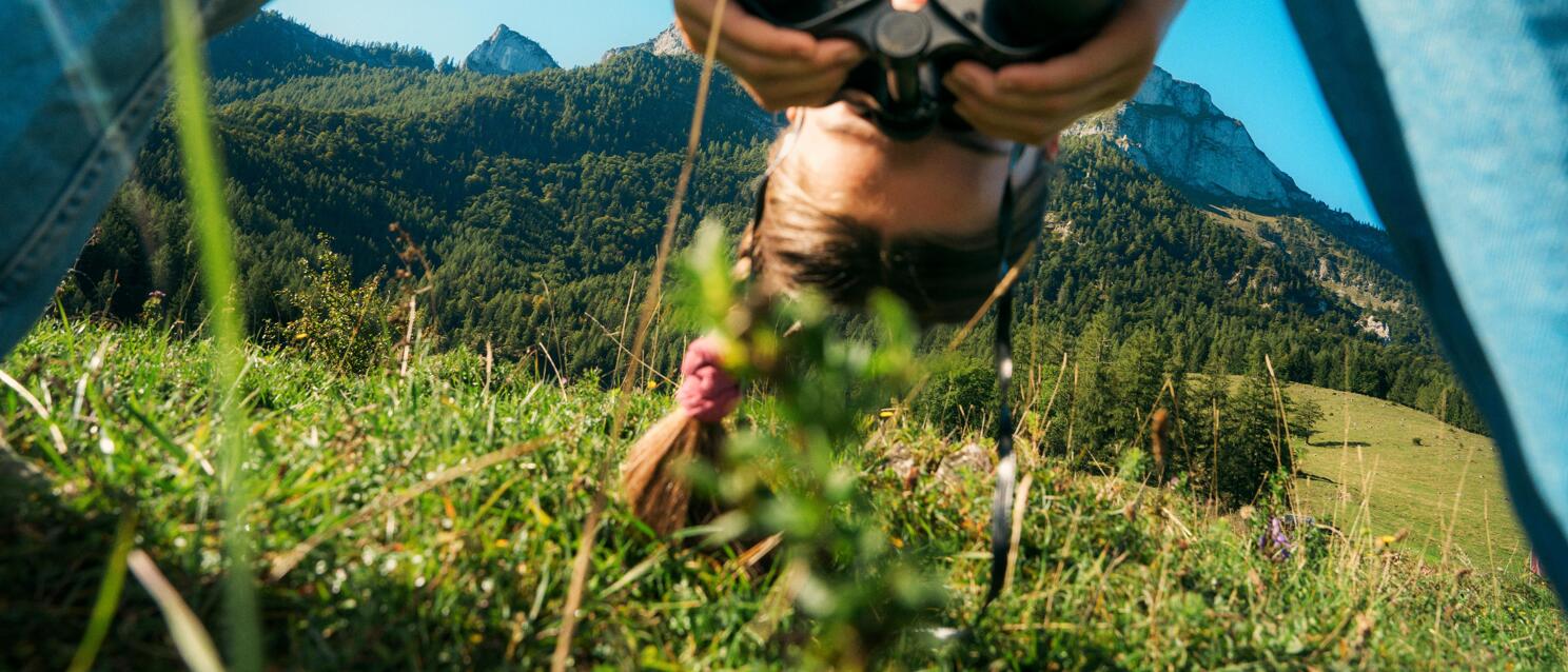 Child wearing straw hat and binoculars on an alpine meadow, mountain panorama with rocky peaks in the background.