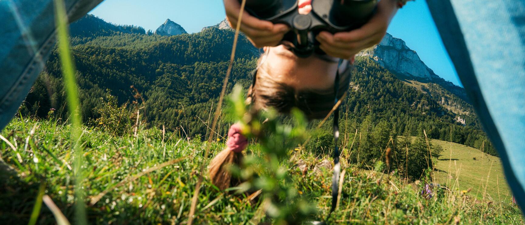 Child wearing straw hat and binoculars on an alpine meadow, mountain panorama with rocky peaks in the background.