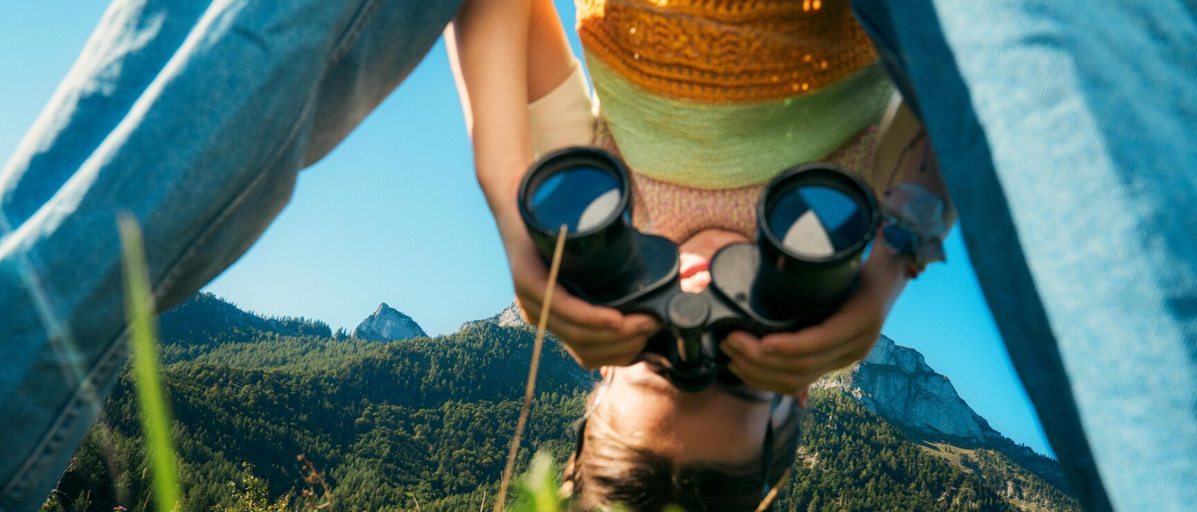 Child wearing straw hat and binoculars on an alpine meadow, mountain panorama with rocky peaks in the background.