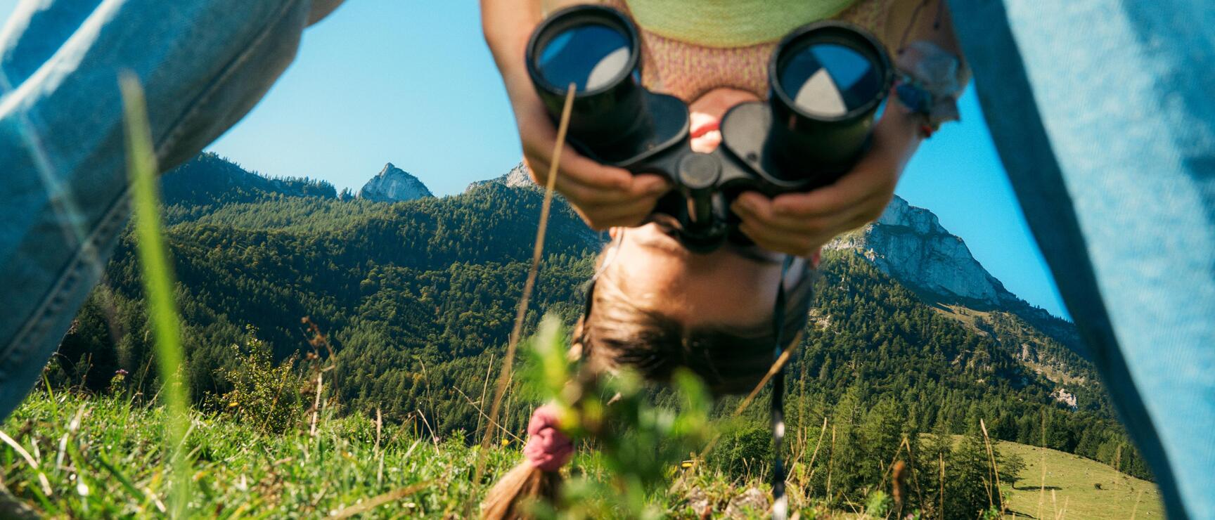 Child wearing straw hat and binoculars on an alpine meadow, mountain panorama with rocky peaks in the background.