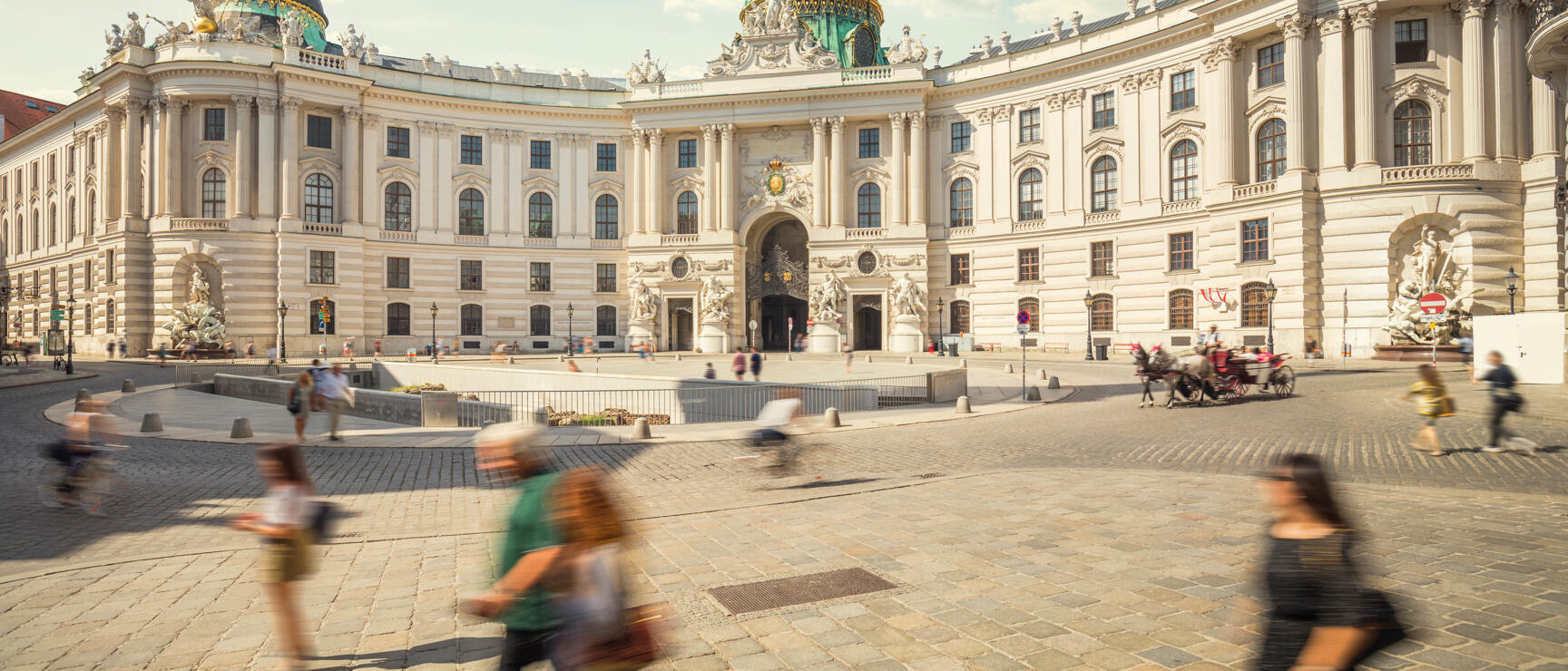 Michaelerplatz with Hofburg, green cupolas and sculptures, pedestrians and fiaker carriages in foreground, Vienna.