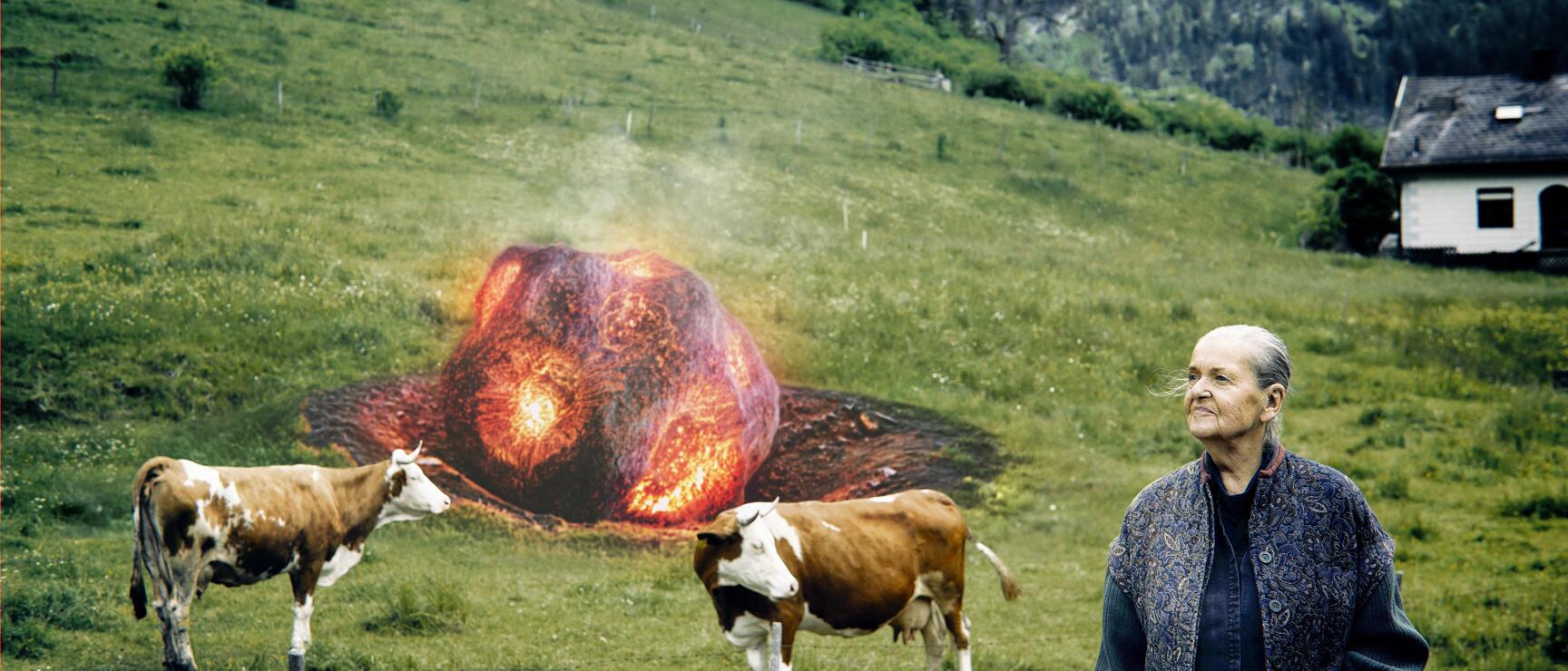 Ältere Frau und zwei Kühe auf grüner Alpenwiese, dahinter digital eingefügter glühender Feuerball.