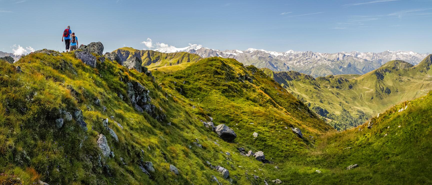 Two hikers on a mountain ridge, green alpine meadows and snow-capped peaks in the background.