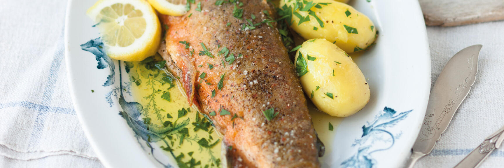 Fried trout with lemon slices and parsley potatoes on white plate with blue pattern.