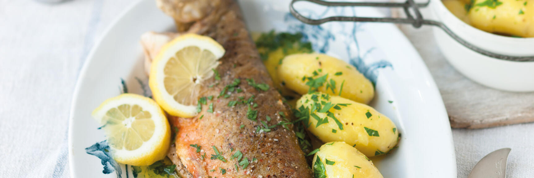 Fried trout with lemon slices and parsley potatoes on white plate with blue pattern.