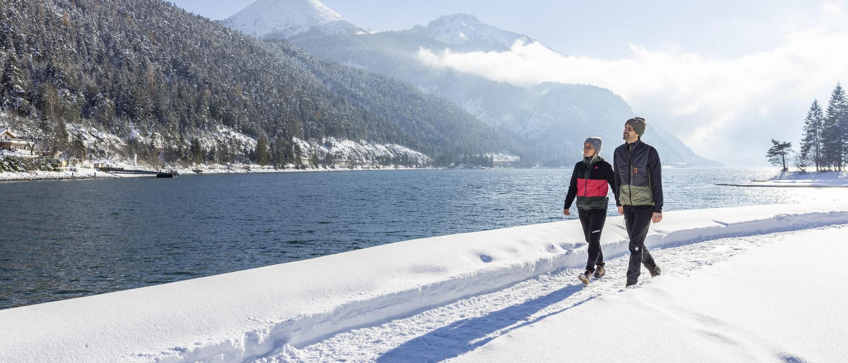 Two people strolling on snow-covered lakeside path, snow-laden forest in background.