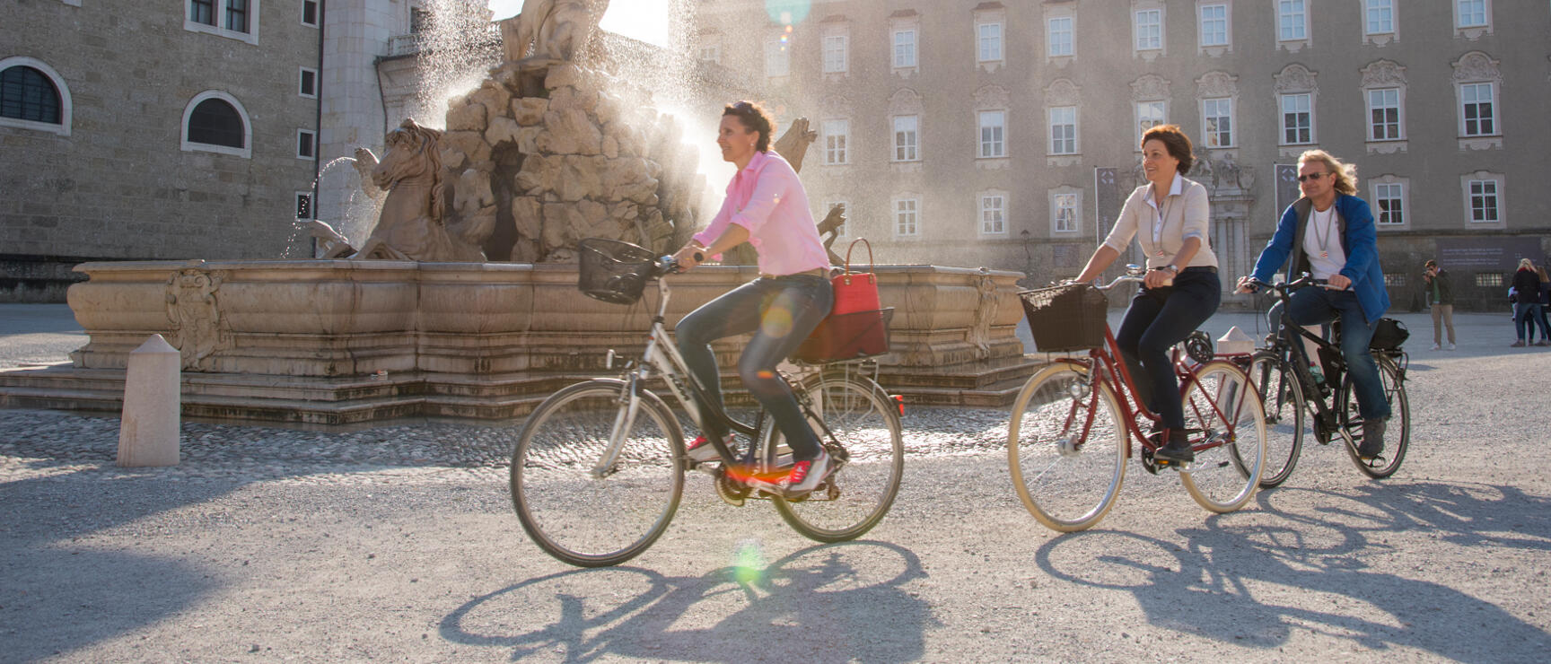 Three women on bicycles at Residenzplatz in Salzburg, baroque fountain and Residenz in the background.