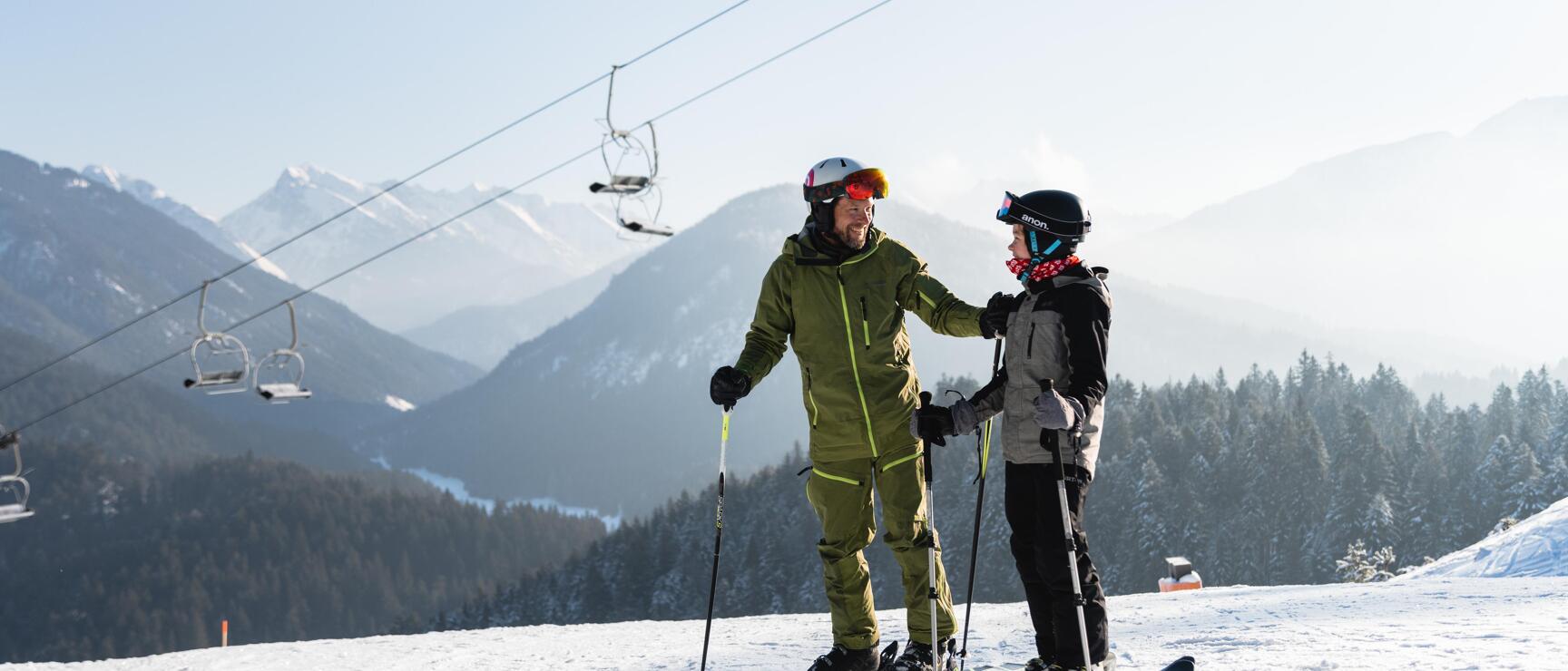 Two skiers on snowy slope under chairlift, mountain panorama with coniferous forest in background.