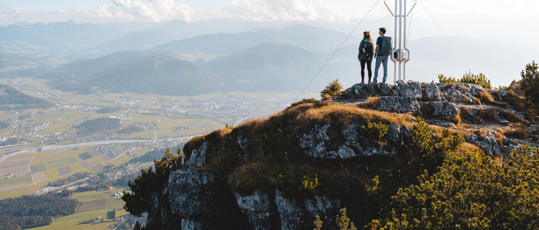 Two hikers with backpacks beside a summit cross on rocks, wide alpine valley in the background.