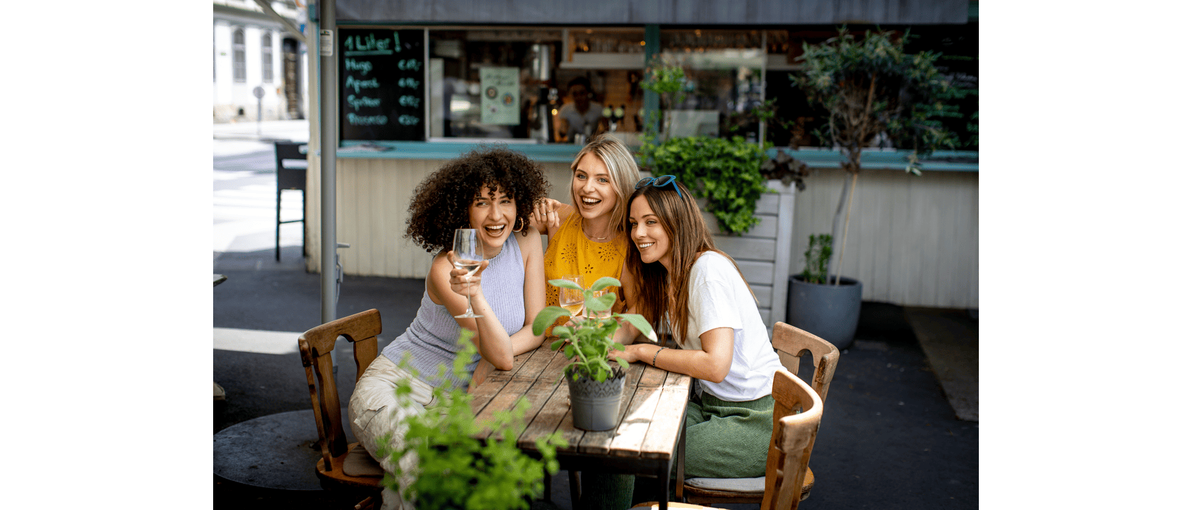 Three laughing women with wine glasses at a wooden table in a garden café, drinks table visible in the background.