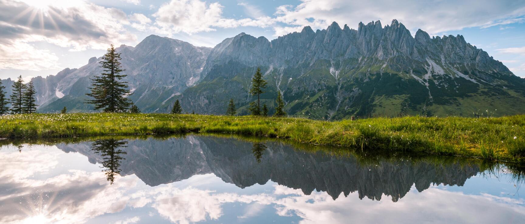 Rocky mountain range with fir trees and green meadow reflected in an alpine lake, sun flare in upper left.