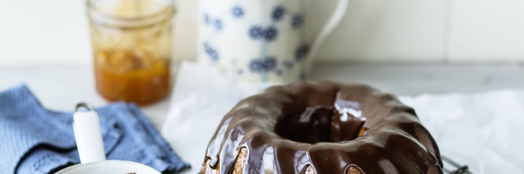 Bundt cake with chocolate glaze on cake rack, beside it bowl with chocolate and pastry brush.