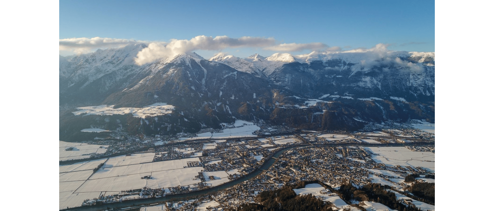 Aerial view of snowy winter town in valley with river, snow-covered fields and mountain range in background.
