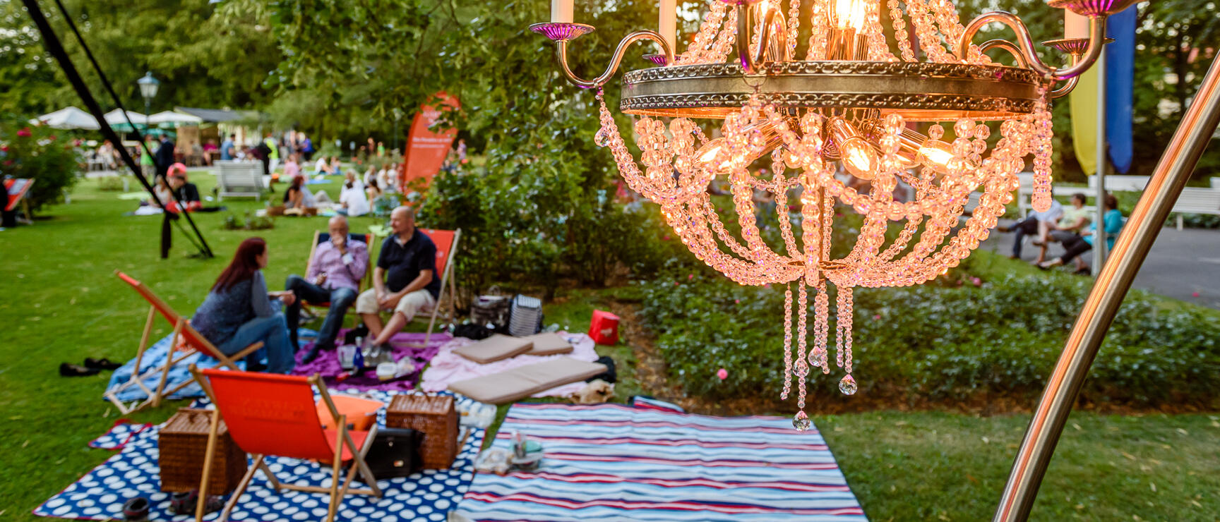 Glowing crystal chandelier outdoors, people on blankets and loungers in a park behind it.