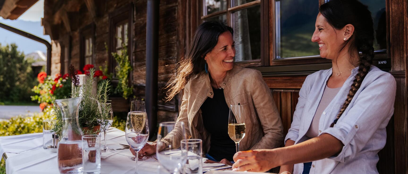 Two women with wine glasses at a set table on a wooden house terrace, red geraniums in the background.