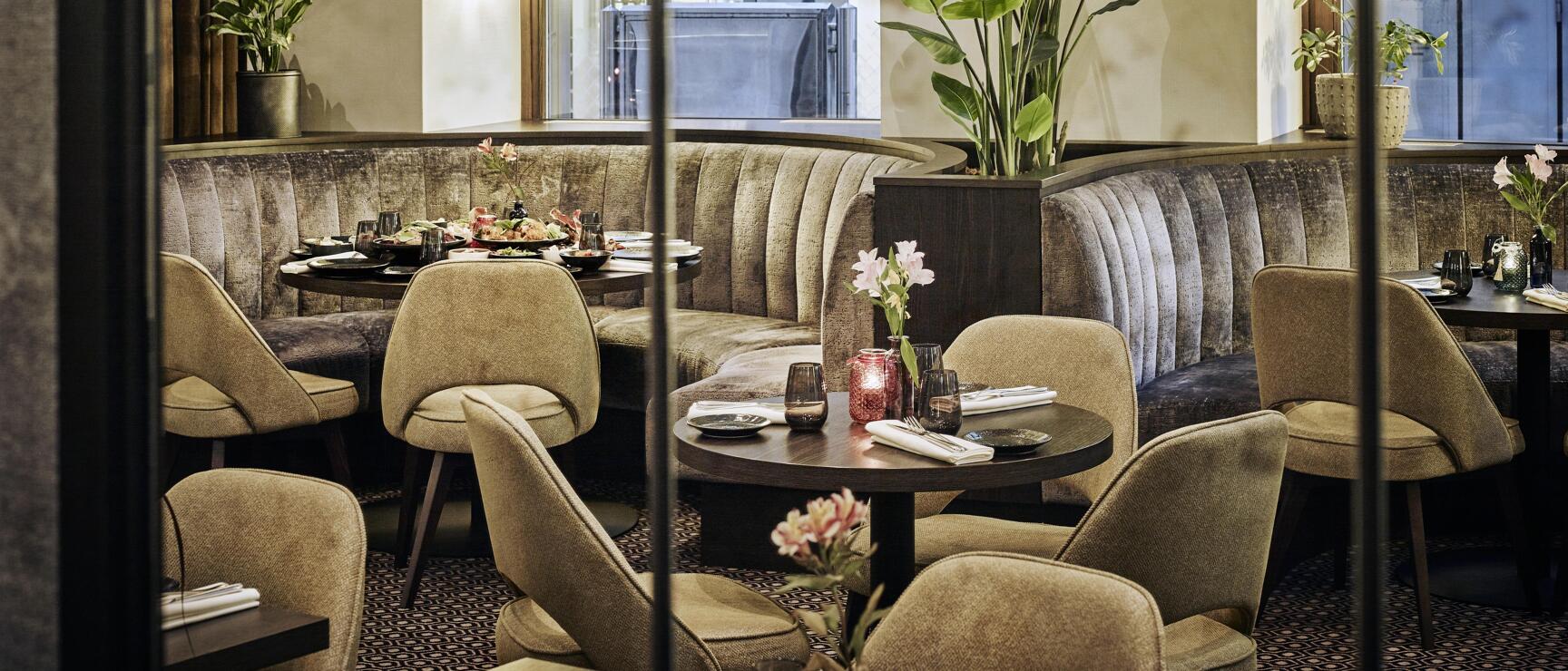 Elegant restaurant interior with velvet upholstery, round tables, candles, green plant and chandelier.