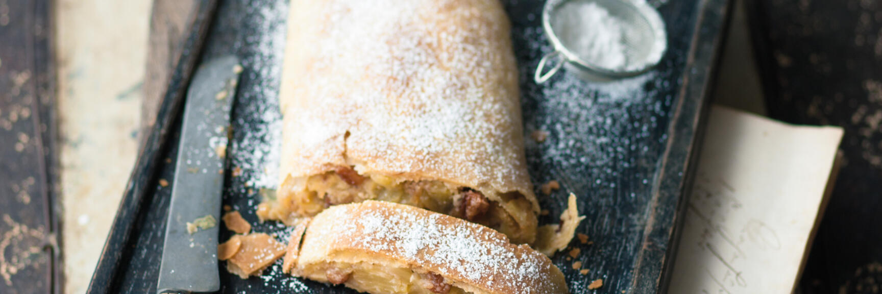 Angeschnittener Apfelstrudel mit Puderzucker auf dunklem Holzbrett, Messer und Puderzuckersieb daneben.