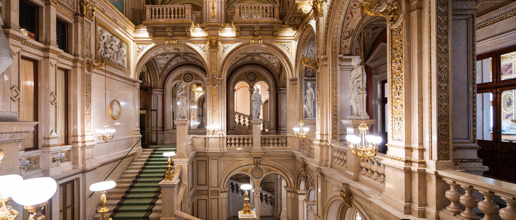 Ornate foyer of the Vienna State Opera with green staircase, marble columns, gold decorations and statues.
