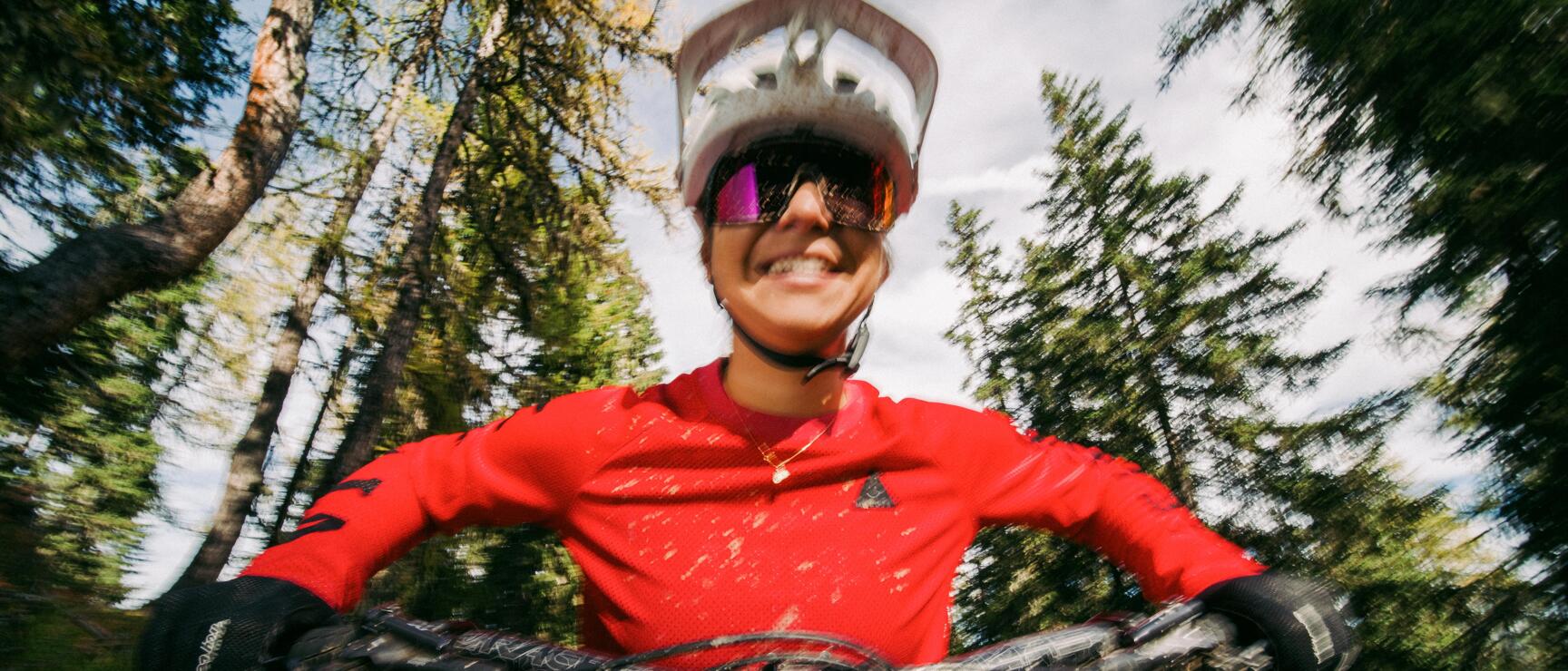 Female mountain biker in white helmet and red jersey smiling while riding through a coniferous forest.