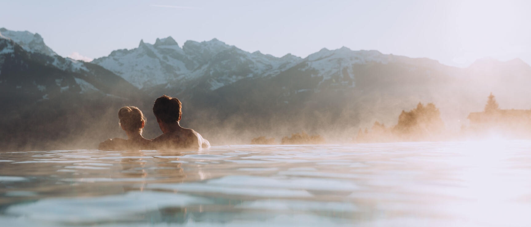 Two people in a steaming outdoor pool overlooking snow-covered mountain peaks.