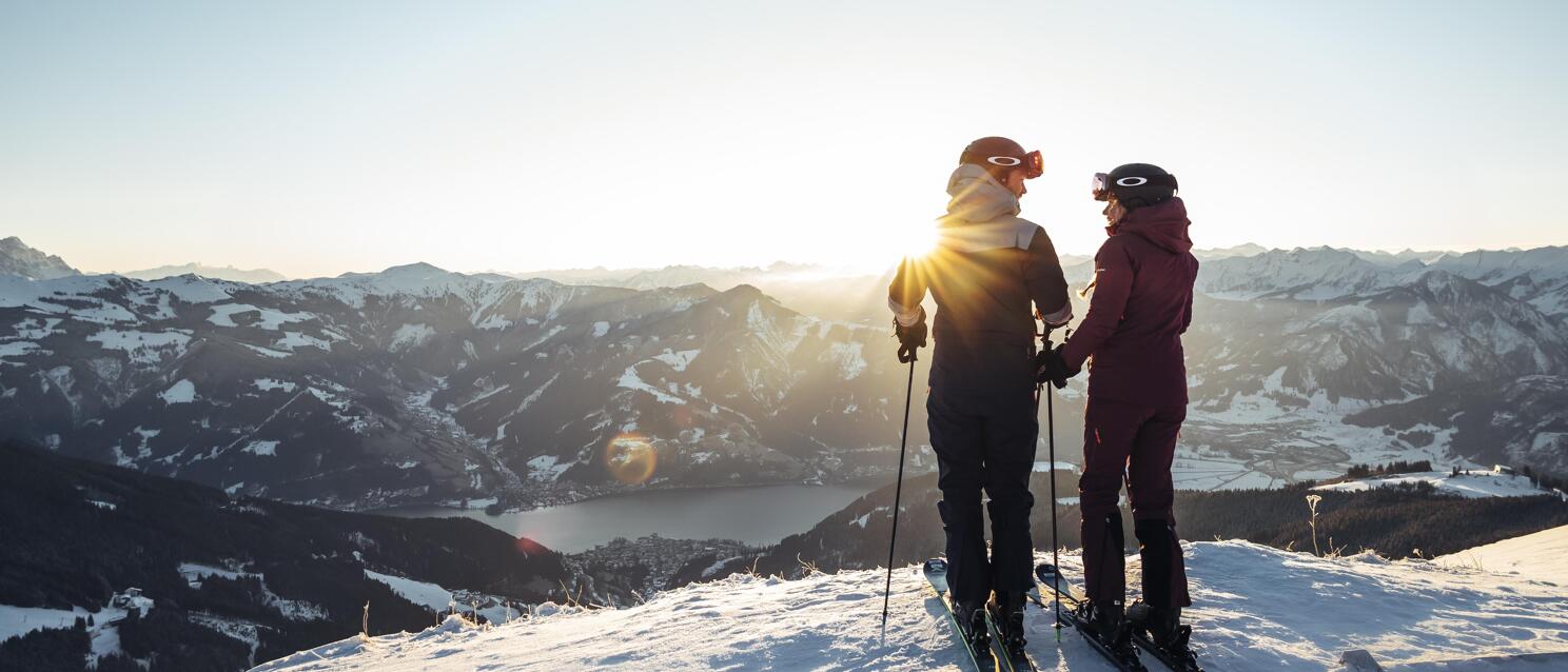 Two skiers on a summit overlooking Lake Zell and snow-covered mountains at sunset.