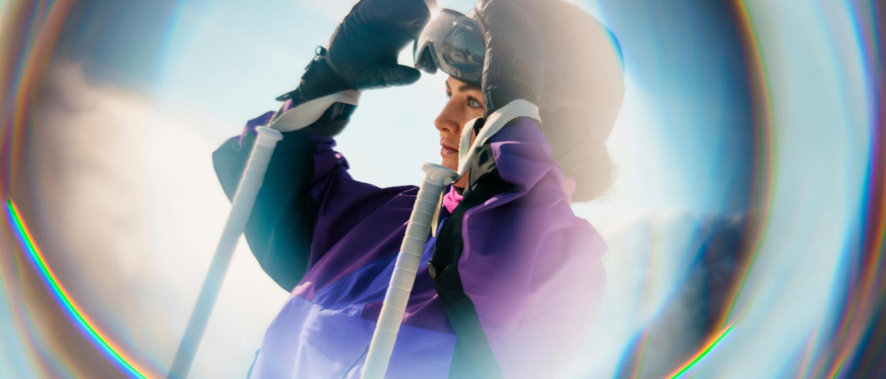 A female skier in purple winter gear adjusts her goggles, while the bright winter light and snow-covered mountain backdrop create a serene alpine atmosphere.