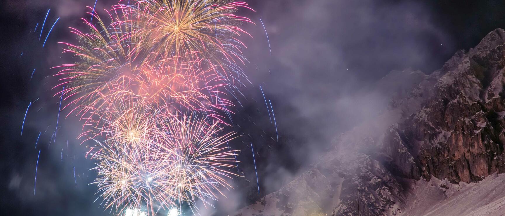 Pink and gold fireworks over snow-covered mountain slope at night with illuminated building on right.