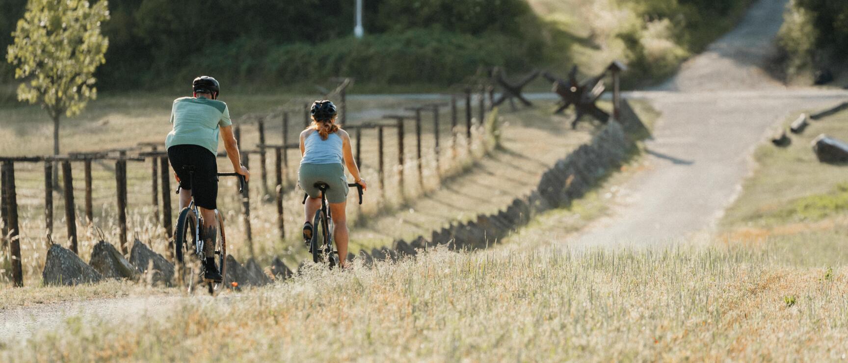 Two cyclists with helmets on a gravel path beside wooden fence and meadow, trees in the background.