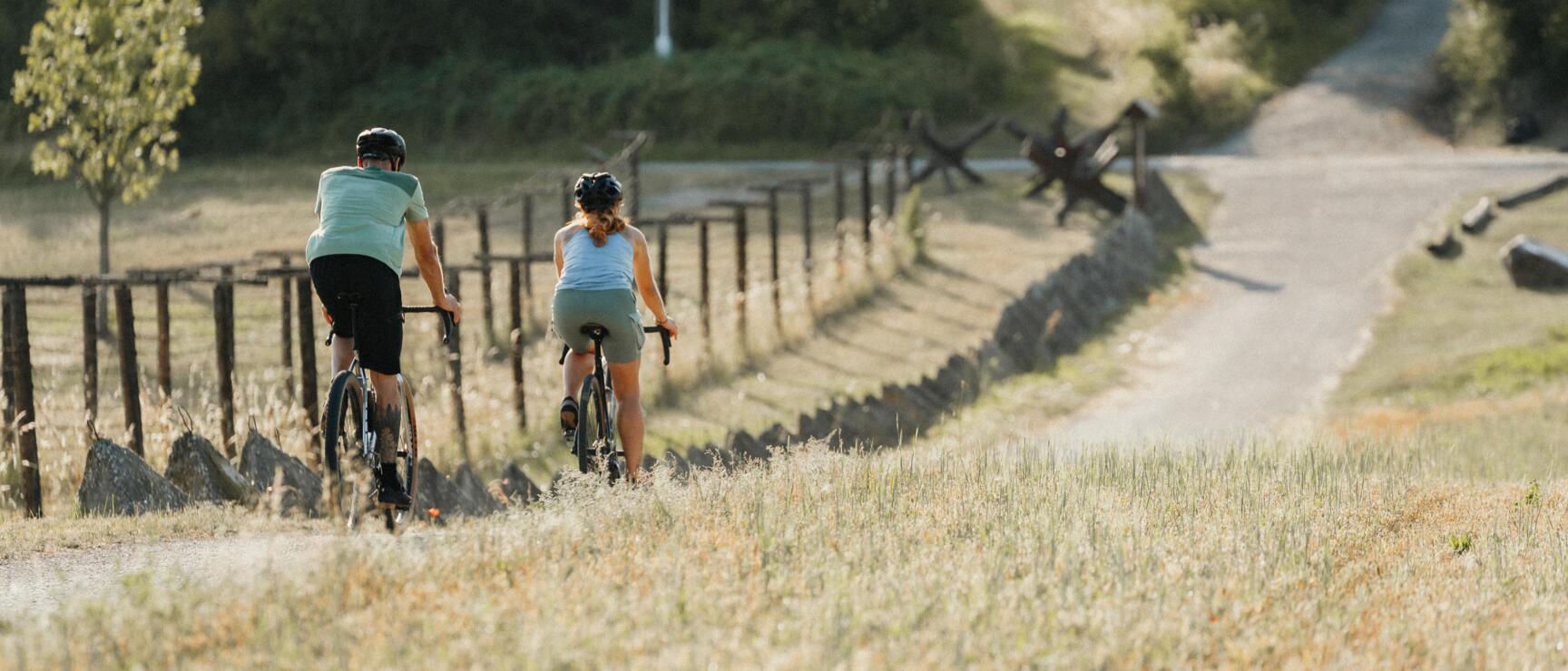 Zwei Radfahrer mit Helmen auf einem Schotterweg neben Holzzaun und Wiese, Bäume im Hintergrund.