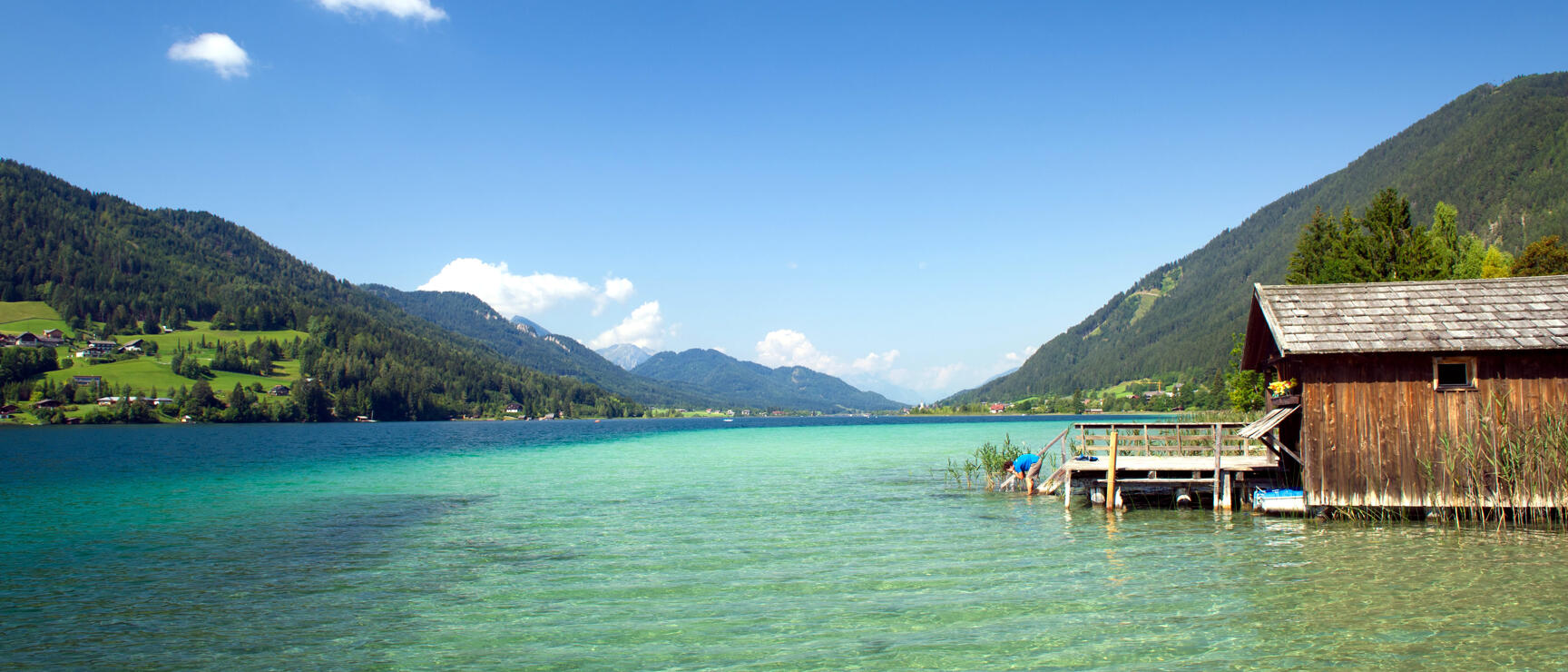 Turquoise mountain lake with wooden boathouse on stilts, forested mountains in the background, blue sky.