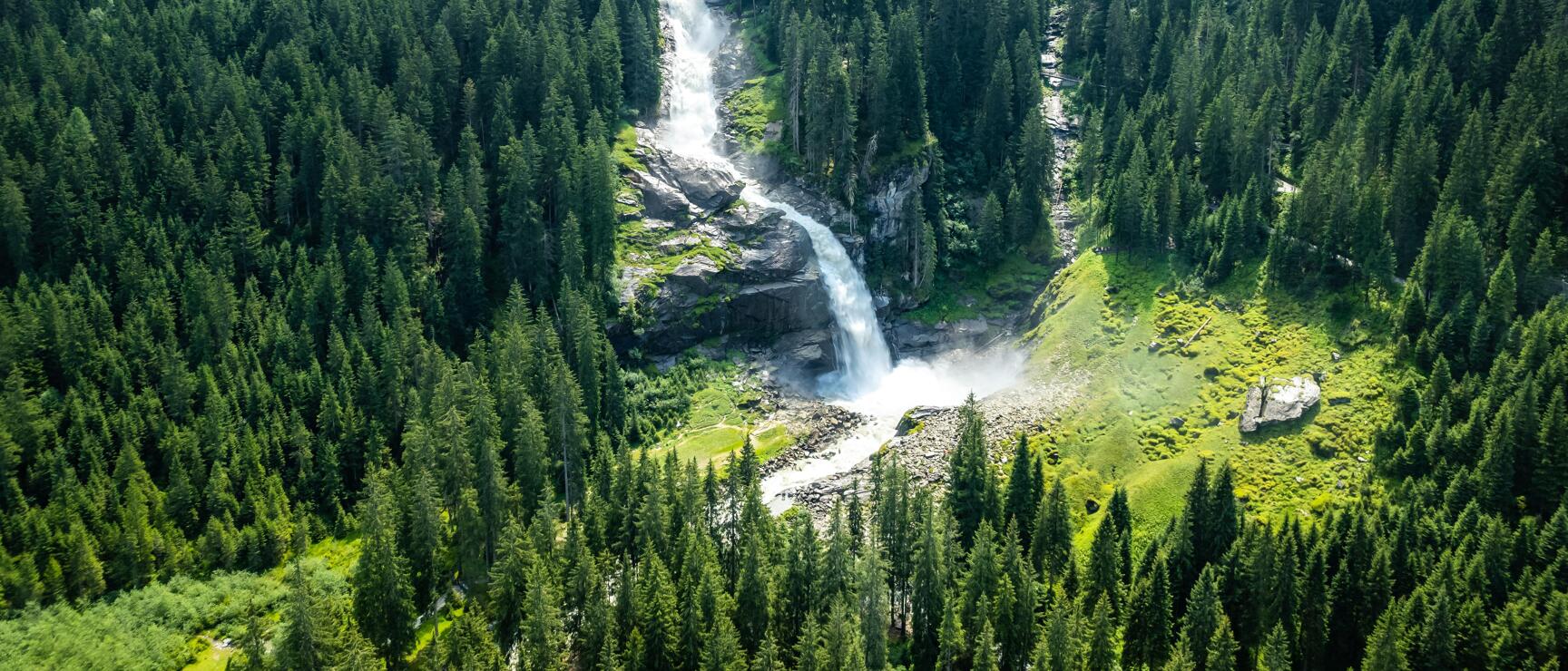Aerial view of multi-tiered waterfall amid dense coniferous forest, mountain valley in the background.